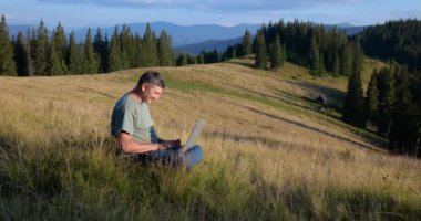 A man sits on a beautiful meadow in the mountains, works on a laptop. Concept of freelancing, digital nomad or remote office.