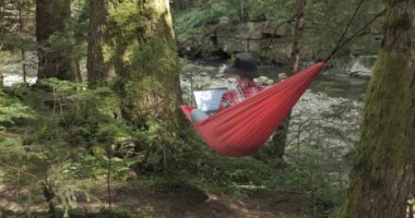 A woman works on a laptop in a hammock on the river bank in a beautiful forest. Concept of freelancing, digital nomad or remote office. 4K