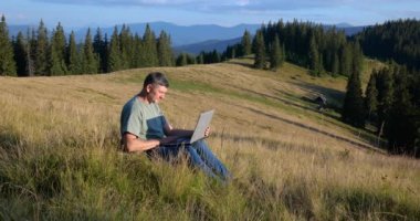 A man sits on a beautiful meadow in the mountains, works on a laptop. Concept of freelancing, digital nomad or remote office.