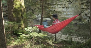 A woman works on a laptop in a hammock on the river bank in a beautiful forest. Concept of freelancing, digital nomad or remote office. 4K