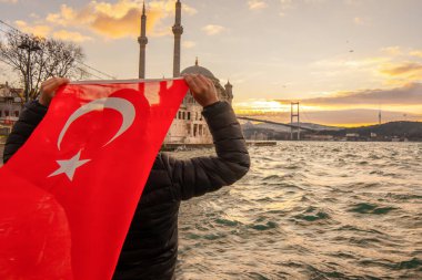 Adam Türk bayrağı taşıyor. İstanbul 'un Ortakoy kentinde Boğaziçi Köprüsü (Türkçe: 15 Temmuz Sehitler Koprusu) ve Sultan Abdulmecid İmparatorluk Camii yakınlarında gün doğumu. Vatanseverlik