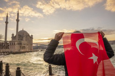 Adam Türk bayrağı taşıyor. İstanbul 'un Ortakoy kentinde Boğaziçi Köprüsü (Türkçe: 15 Temmuz Sehitler Koprusu) ve Sultan Abdulmecid İmparatorluk Camii yakınlarında gün doğumu. Vatanseverlik