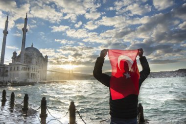 Adam Türk bayrağı taşıyor. İstanbul 'un Ortakoy kentinde Boğaziçi Köprüsü (Türkçe: 15 Temmuz Sehitler Koprusu) ve Sultan Abdulmecid İmparatorluk Camii yakınlarında gün doğumu. Vatanseverlik