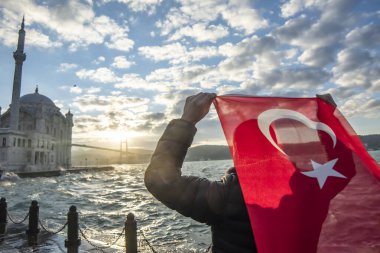 Adam Türk bayrağı taşıyor. İstanbul 'un Ortakoy kentinde Boğaziçi Köprüsü (Türkçe: 15 Temmuz Sehitler Koprusu) ve Sultan Abdulmecid İmparatorluk Camii yakınlarında gün doğumu. Vatanseverlik