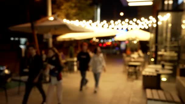 Pessoas desfocadas andando ao longo do bistrô com iluminação noturna embaçada bokeh ao ar livre, beco — Vídeo de Stock