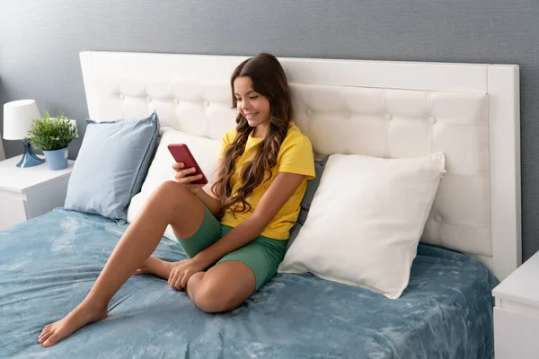 快乐的女孩坐在床上发短信，在手机上发短信 — 图库照片