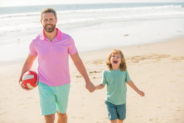 sport activity. smiling father and son walk with ball on beach. daddy with kid boy