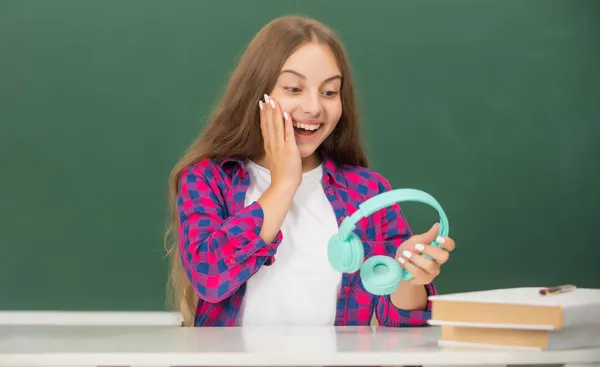 Surpris adolescent fille tenir écouteurs modernes au lycée au tableau noir, école — Photo