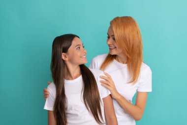Sevgi dolu, gülümseyen bir kadın mavi arka planda beyaz gömlekli bir kıza sarılıyor.
