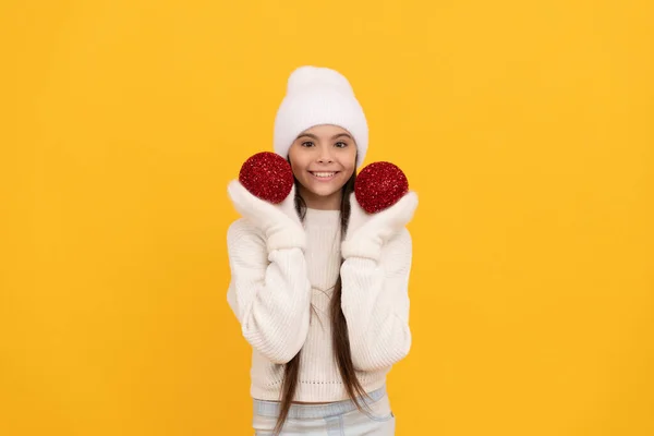 Fröhliches Kind mit Wintermütze und Handschuhen hält dekorative Weihnachtskugeln auf gelbem Hintergrund, Weihnachten — Stockfoto