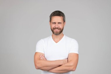 Mutlu, kendine güvenen adam gülümsüyor kollarını beyaz tişörtlü, gri arka planda tutuyor.