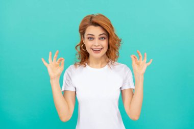 Kıvırcık saçlı, şaşkın bir kızıl kız mavi arka planda güzel bir jest yapıyor.