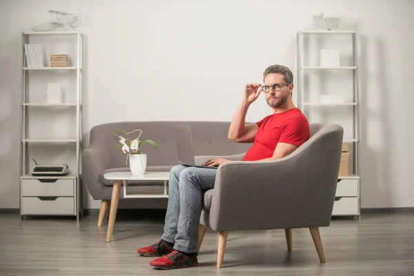 Serious guy in red tshirt and eyeglasses work on pc in chair, home office — Stock Photo, Image