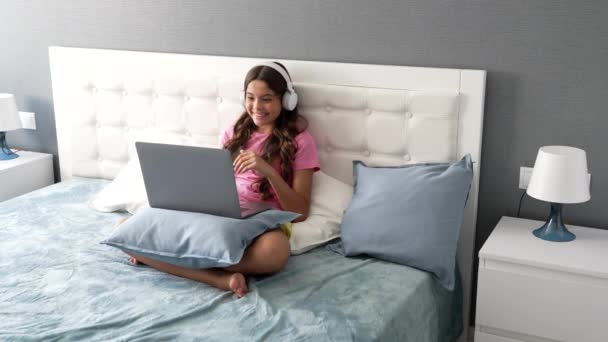 Feliz niña pequeña en los auriculares comenzó a elearning con el ordenador en línea en la cama, la infancia — Vídeo de stock