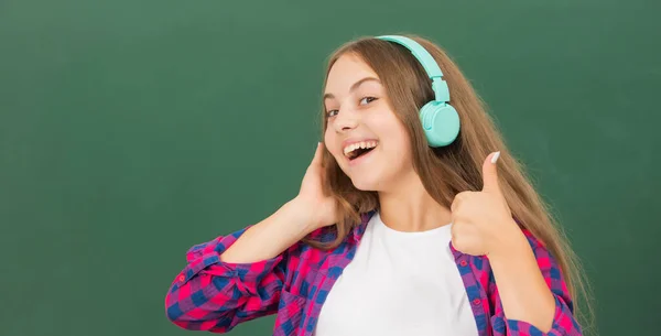 Fille heureuse tenir écouteurs modernes au lycée au tableau noir montrant pouce vers le haut, accessoire audio. — Photo