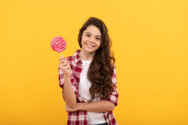 Uzun kıvırcık saçlı mutlu çocuk sarı arka planda lolipop karamel şekeri tutuyor..