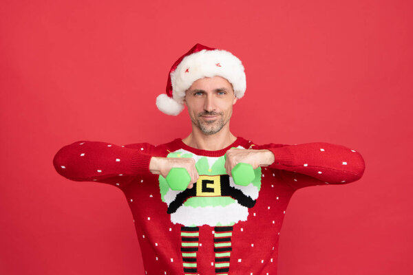 Счастливый Санта-мужчина улыбается, делая гантели тренировки на красном фоне, Рождество
