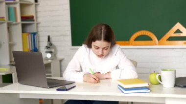 Ciddi bir kız çocuğu okul masasında, okul kitapçığında yazı yazıyor.