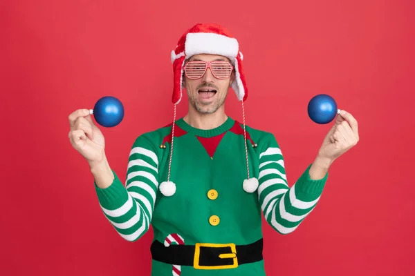 Hombre sorprendido en traje de elfo y sombrero de Santa Claus. chico en gafas de fiesta celebrar bolas de decoración de Navidad —  Fotos de Stock