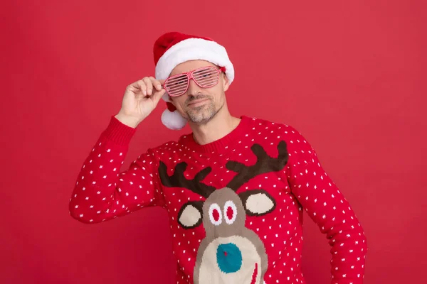 Усміхнений чоловік в зимовому светрі і партійних окулярах. Різдвяний хлопець у капелюсі Санта Клауса на червоному тлі — стокове фото