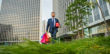 Takım elbiseli güler yüzlü girişimci elinde alışveriş çantası ve hediye kutusuyla ofisin dışında alışveriş yapıyor..