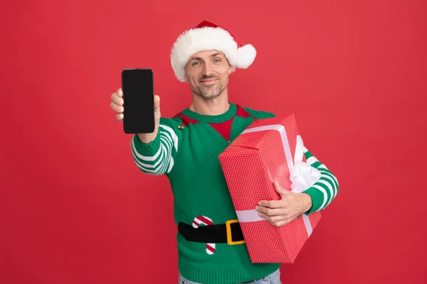 Šťastný elf muž v Santa Claus klobouk prezentující smartphone. vánoční reklama. — Stock fotografie