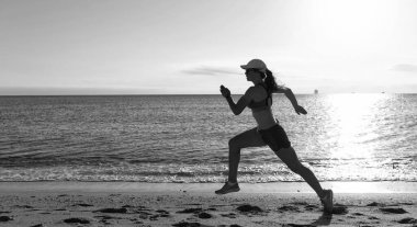 Hareketli giyimli enerjik bir kadın sahil kenarında kumsalda koşuyor.