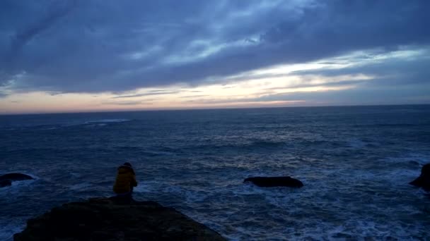 Одинокая девушка в куртке, сидящая у волнистого океана с драматическим небом, думая: — стоковое видео