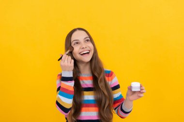 Mutlu çocuk makyaj malzemelerini pudra fırçası sarı arka plan ve makyajla kullanıyor.