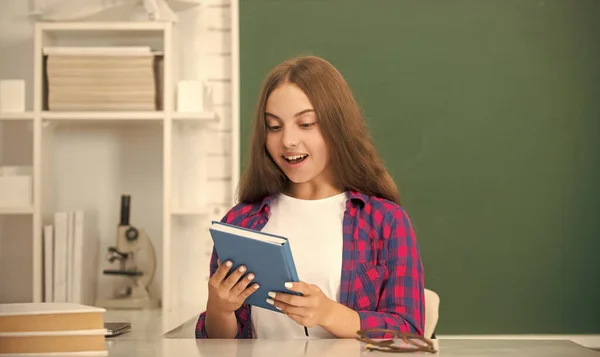 Amazed kid sitting with copybook in classroom at blackboard, education — Stock Photo, Image