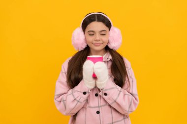 teen girl in earmuffs smell coffee or tea cup on yellow background, morning