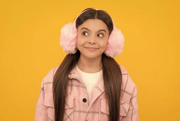 Happy girl child in warm winter fur earmuffs smile with sly look yellow background, intrigue — 스톡 사진