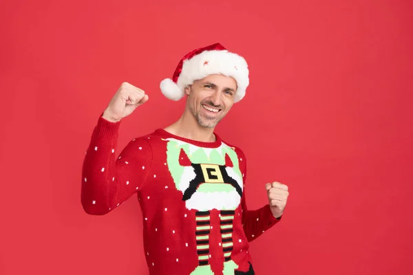 Happy guy in Santa Claus hat and Christmas jumper make winning gesture red background, new year — стоковое фото