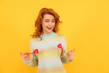 happy cheerful redhead girl with red heart sticks on yellow background. february 14