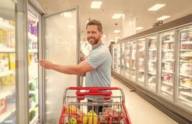 Alışveriş arabalı mutlu bir adam marketten yiyecek alıyor, alışveriş yapıyor.