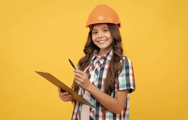 Cheerful kid in construction helmet making notes in clipboard, taking notes — Zdjęcie stockowe