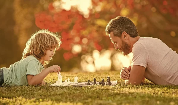 Família feliz de pai homem e filho criança jogando xadrez na grama verde no parque ao ar livre, paternidade — Fotografia de Stock