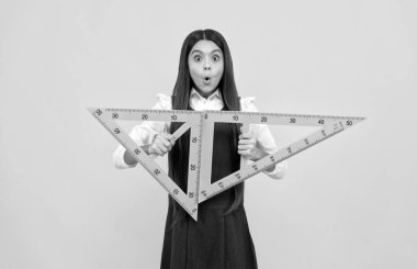 Şaşırmış genç kız okulda sarı arka planda matematik üçgeni cetvelini tutuyor.