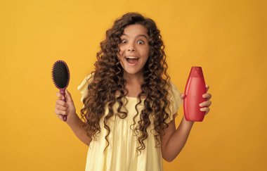 Şok olmuş kıvırcık saçlı çocuk tarak ve şampuan şişesini tutuyor. Genç güzel saç stili.