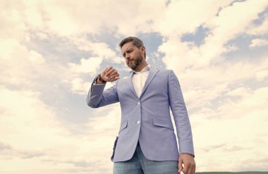 Resmi takım elbiseli yakışıklı, olgun iş adamı. Kol saatini kontrol et. Gökyüzü arka planında, zamanda.