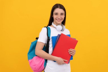 Sırt çantası ve kulaklıklı genç kız sarı arka planda çalışma kitabını tutuyor, okula geri dönüyor.