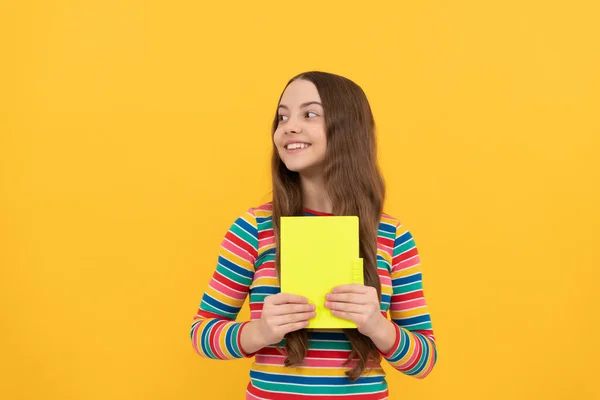 Счастливая девочка улыбается, держа книгу на желтом фоне, школа — стоковое фото