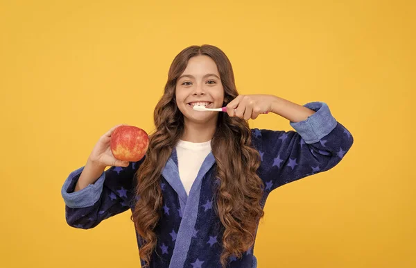 Vrolijk tiener meisje met krullend haar in comfortabele pyjama poetsen tanden en houden appel, tandheelkunde — Stockfoto
