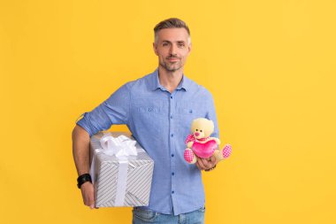 Gülümseyen adam hediye kutusunu ve oyuncağı sarı arka planda tut, doğum günü