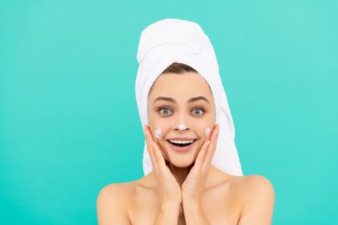 Şaşırmış genç bir kadın mavi arka plana yüz kremi sürmüş.