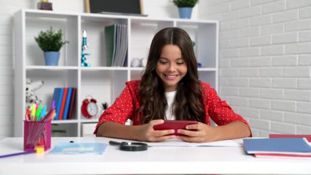 Boldog lány gyermek játszani mobiltelefon játékok iskolai asztalnál, okostelefon — Stock videók