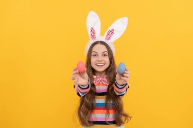 Tavşan kulaklı ve papyonlu neşeli çocuk sarı arka planda Paskalya yumurtası tutuyor.