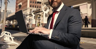 Kırpılmış işadamı kentte bankta oturuyor. Bilgisayarda ve iş bloglarında çalışıyor..