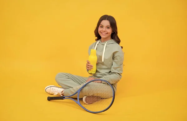 Criança feliz sentar em sportswear com garrafa de água raquete de tênis no fundo amarelo, equilíbrio de água — Fotografia de Stock