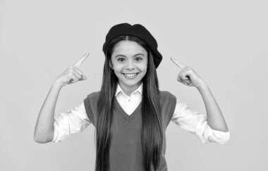 Mutlu liseli kız sarı arka planda Fransız beresini işaret ediyor.